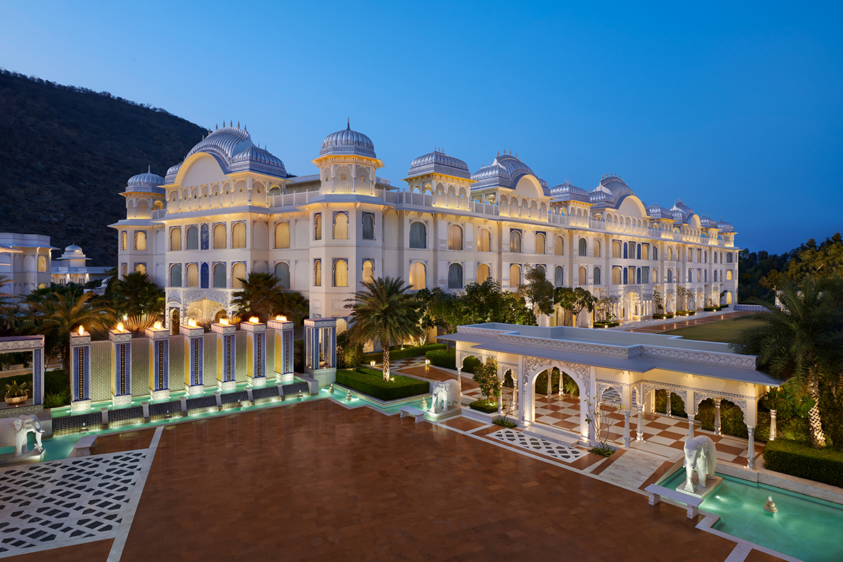 A large, white palace hotel in India along the Leela Palce Trail, with very ornate architecture, beautiful landscape and lit up exterior hotel grounds at night.